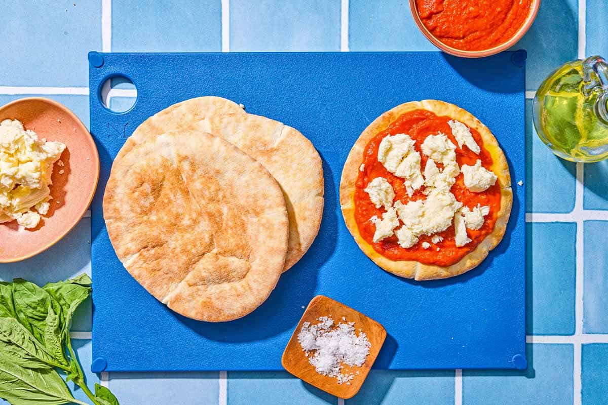A cutting board with a unbaked pita pizza, a stack of 2 pitas and a small bowl of salt. Next to this is a bottle of olive oil, a bowl of marinara, fresh basil leaves and a bowl of fresh mozzarella.