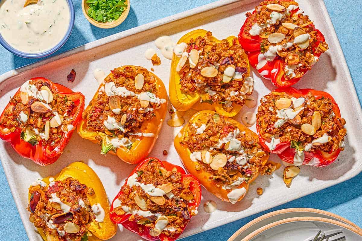 The quinoa stuffed peppers drizzled with tahini sauce in a baking dish. Next to this is a bowl of tahini sauce with a spoon, a bowl of chopped parsley, and a stack of plates.