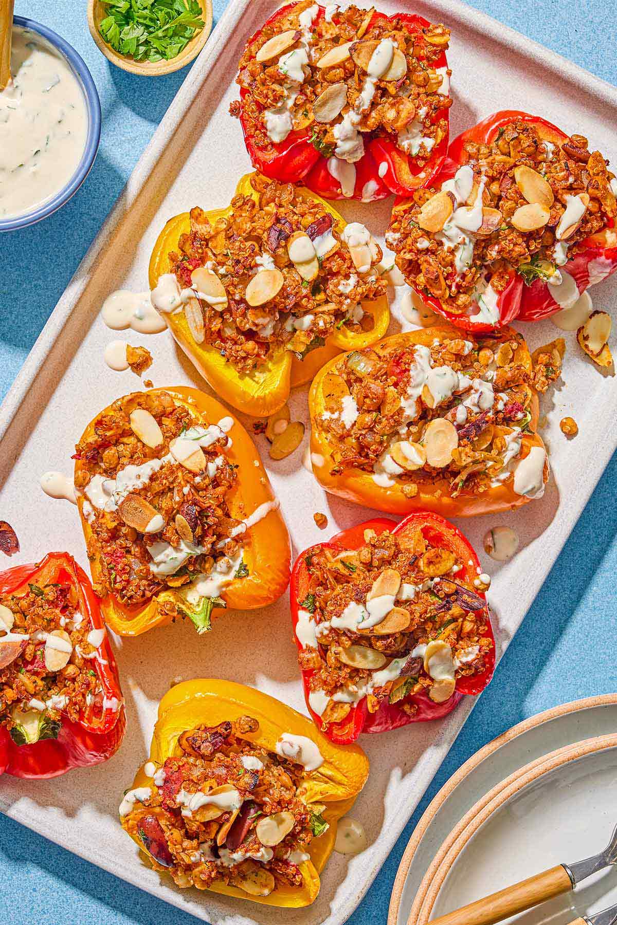 The quinoa stuffed peppers drizzled with tahini sauce in a baking dish. Next to this is a bowl of tahini sauce with a spoon, a bowl of chopped parsley, and a stack of plates with utensils.