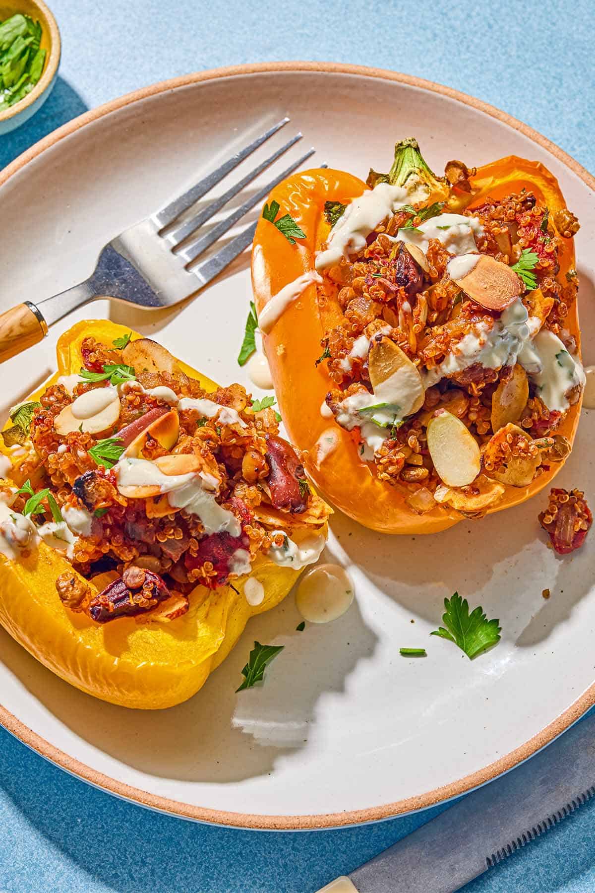 Two quinoa stuffed peppers drizzled with tahini sauce on a plate with a fork. Next to this is a knife and a small bowl of chopped parsley.