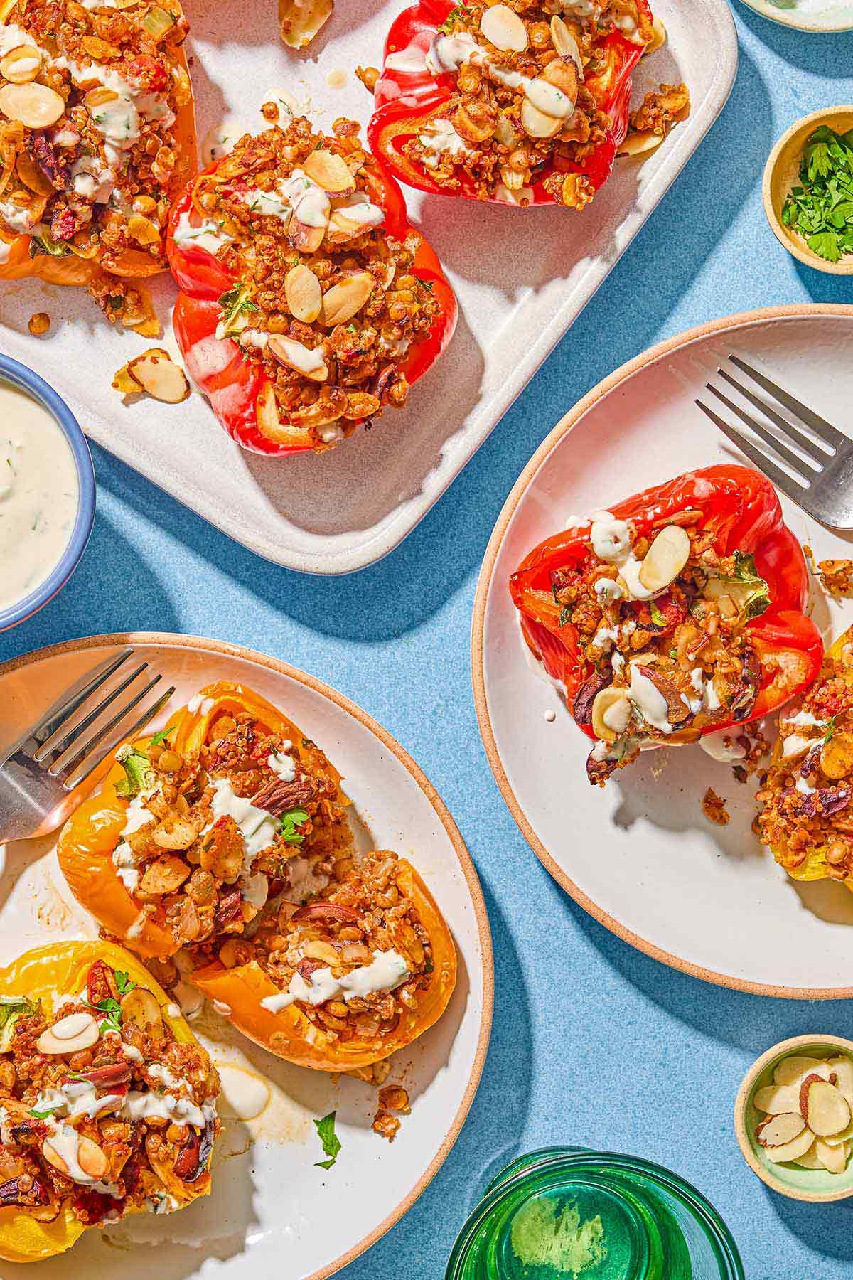 Two servings of the quinoa stuffed peppers on plates with forks. Next to these are bowls of tahini sauce, chopped parsley and almonds and a glass of water.