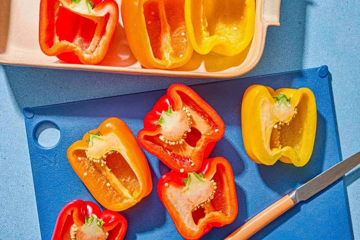 5 bell pepper halves with their seeds removed on a cutting board with a knife. Next to this is a baking dish with 3 more seeded bell pepper halves.