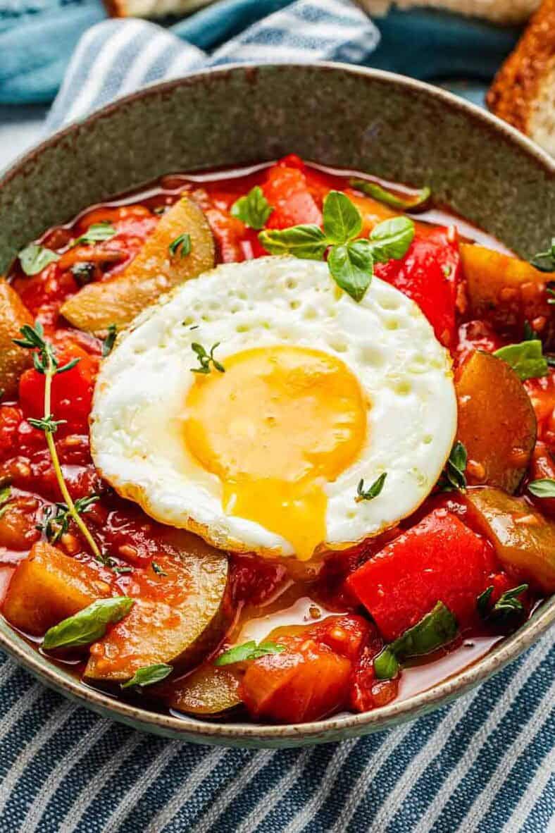 ratatouille topped with a fried egg in a bowl next to a spoon, and slices of toasted bread.