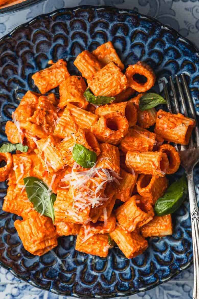 a blue plate with creamy roasted red pepper pasta topped with parmesan cheese and basil with a fork next to a pot of roasted red pepper pasta.