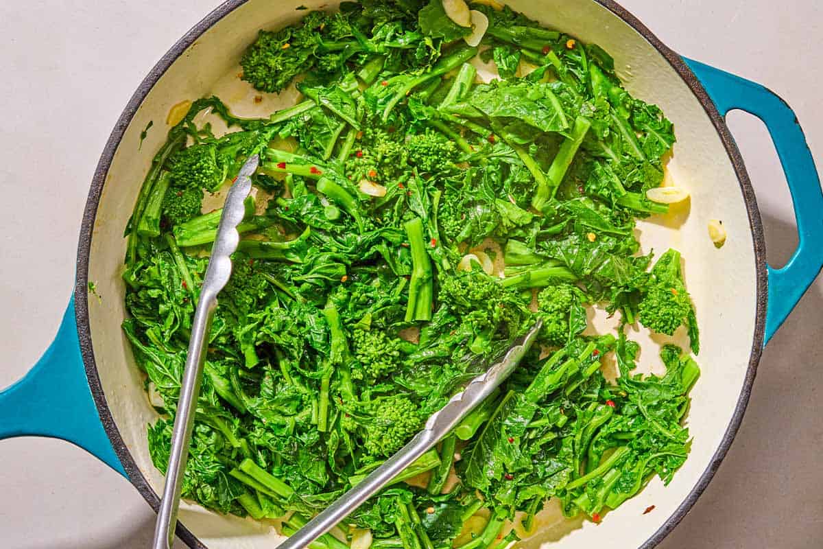 Broccoli rabe being cooked in a skillet with a pair of tongs.