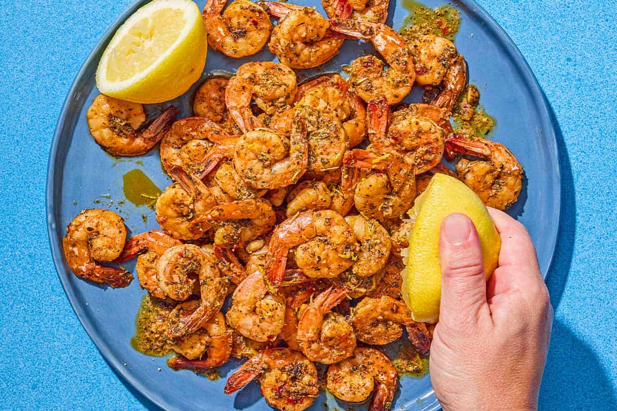 A lemon half being squeezed over the sautéed shrimp that is on a platter with another lemon half.