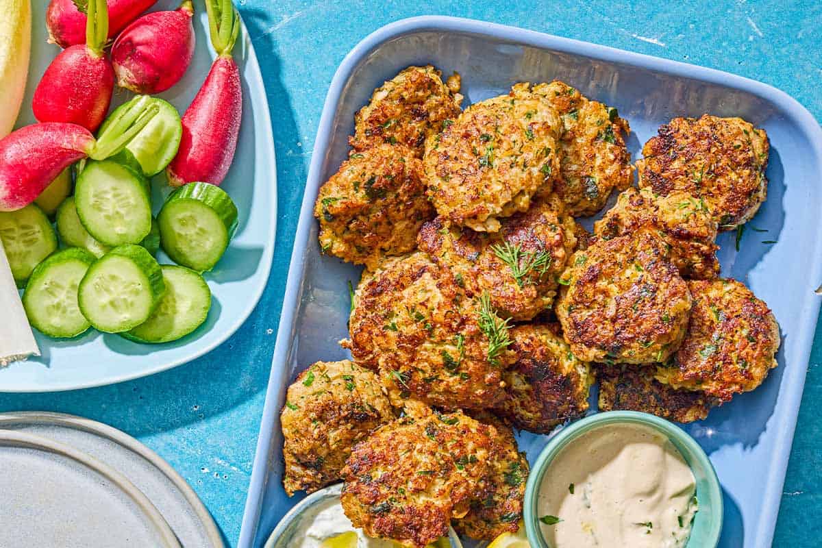 Shrimp kofta on a platter with bowls of tahini and tzaziki sauce and lemon wedges. Next to this is another platter with sliced cucumbers and radishes, and a stack of two plates.