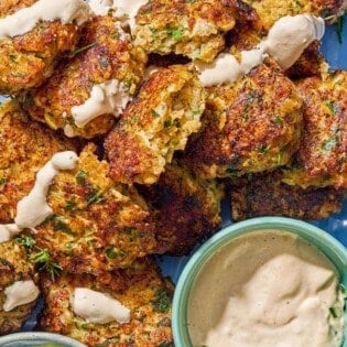 Shrimp kofta drizzled with tahini sauce on a platter with bowls of tahini and tzaziki sauce and lemon wedges.