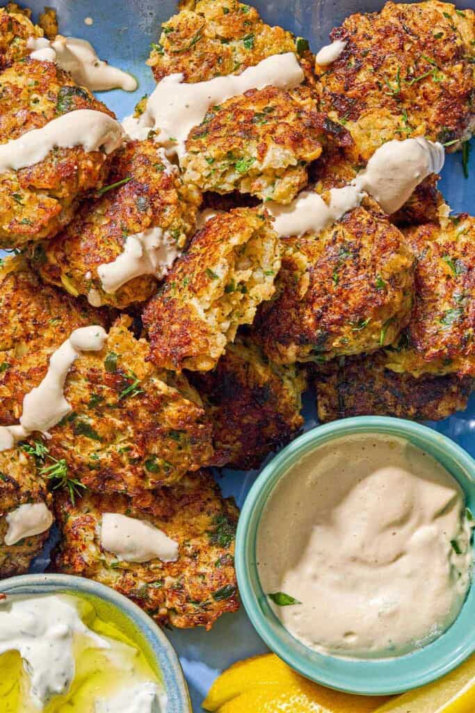 Shrimp kofta drizzled with tahini sauce on a platter with bowls of tahini and tzaziki sauce and lemon wedges.