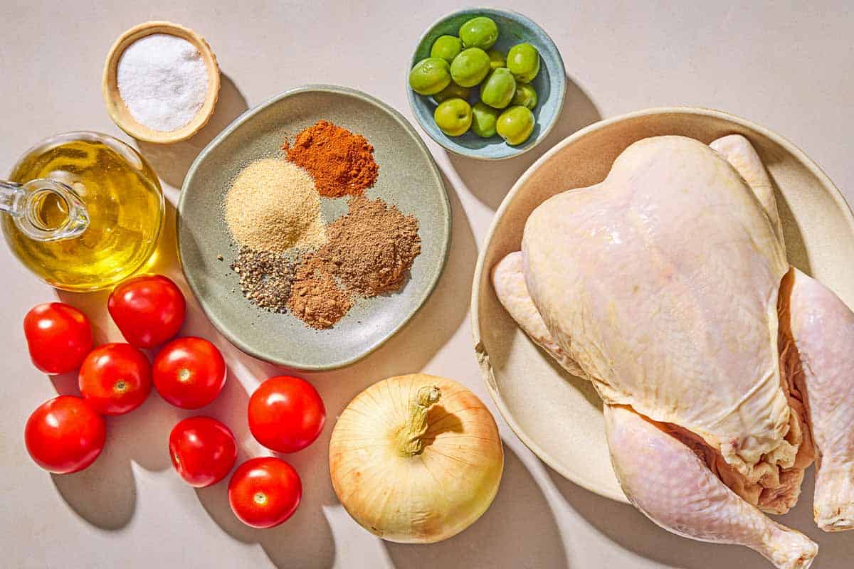 Ingredients for spatchcock chicken including a whole chicken, onion, tomatoes, green olives, olive oil, salt, allspice, garlic powder, sweet paprika, pepper, and nutmeg.