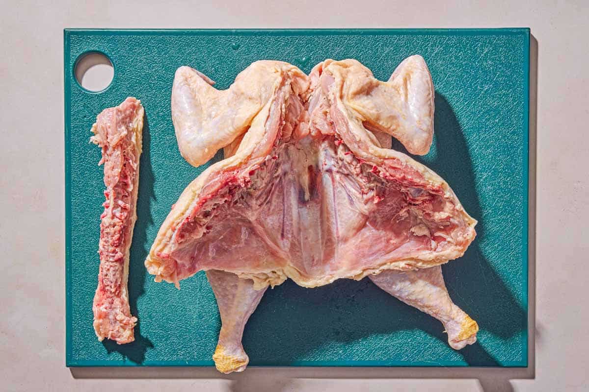 A whole raw chicken with its backbone removed next to its backbone on a cutting board.