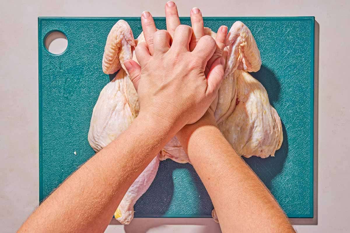 The raw whole chicken being pressed down on a cutting board to flatten.