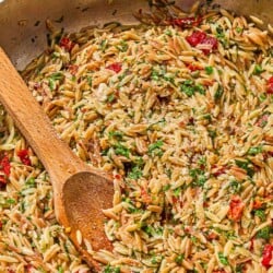The orzo recipe in a skillet with a wooden spoon. Next to this are bowls of grated parmesan cheese and red pepper flakes.