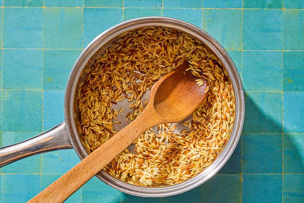 The orzo for the orzo recipe being toasted in a saucepan with a wooden spoon.