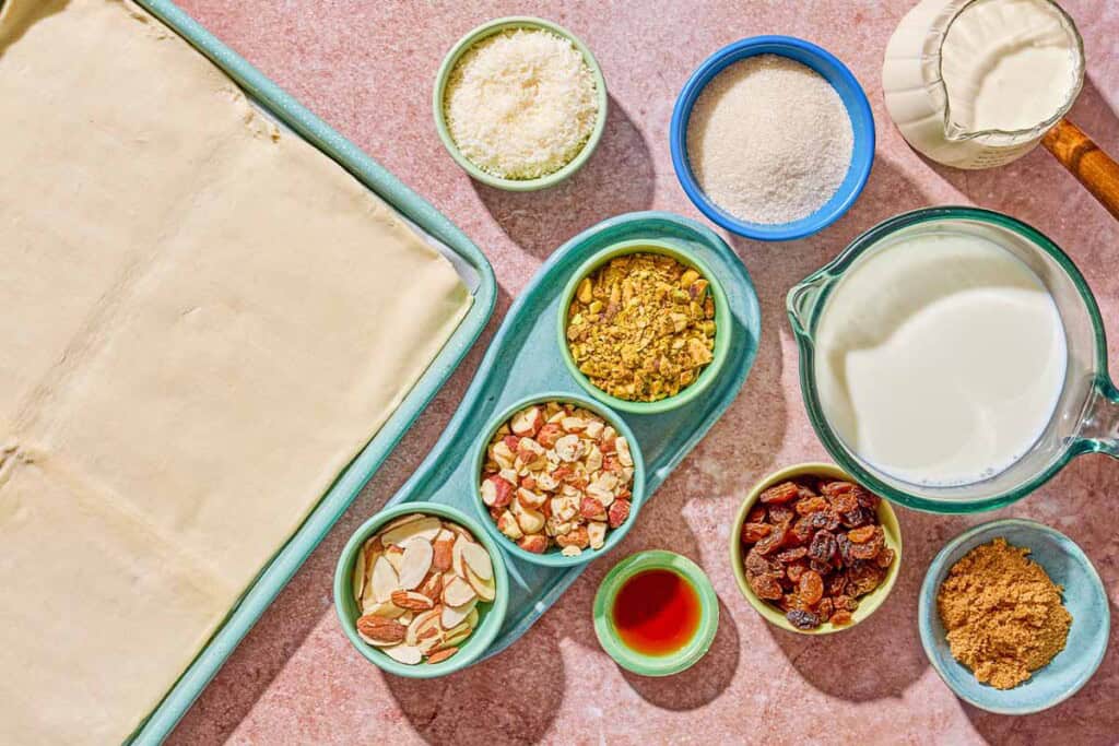 Ingredients for bread pudding including puff pastry, hazelnuts, almonds, pistachios, shredded coconut, raisins, milk, sugar, vanilla extract, heavy cream, brown sugar.