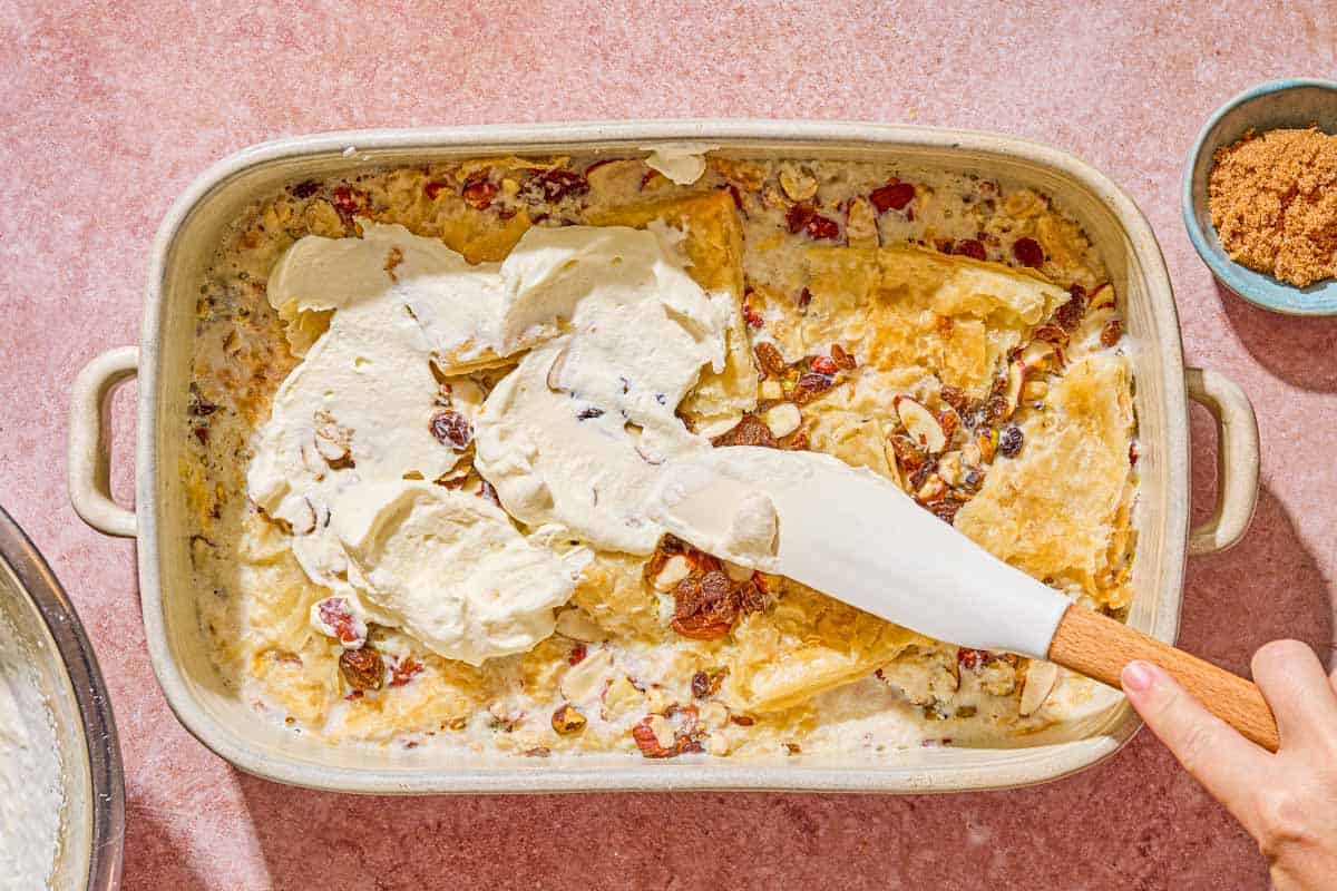 The whipped cream being spread on the unbaked bread pudding in the baking dish. next to this is dark brown sugar in a bowl.