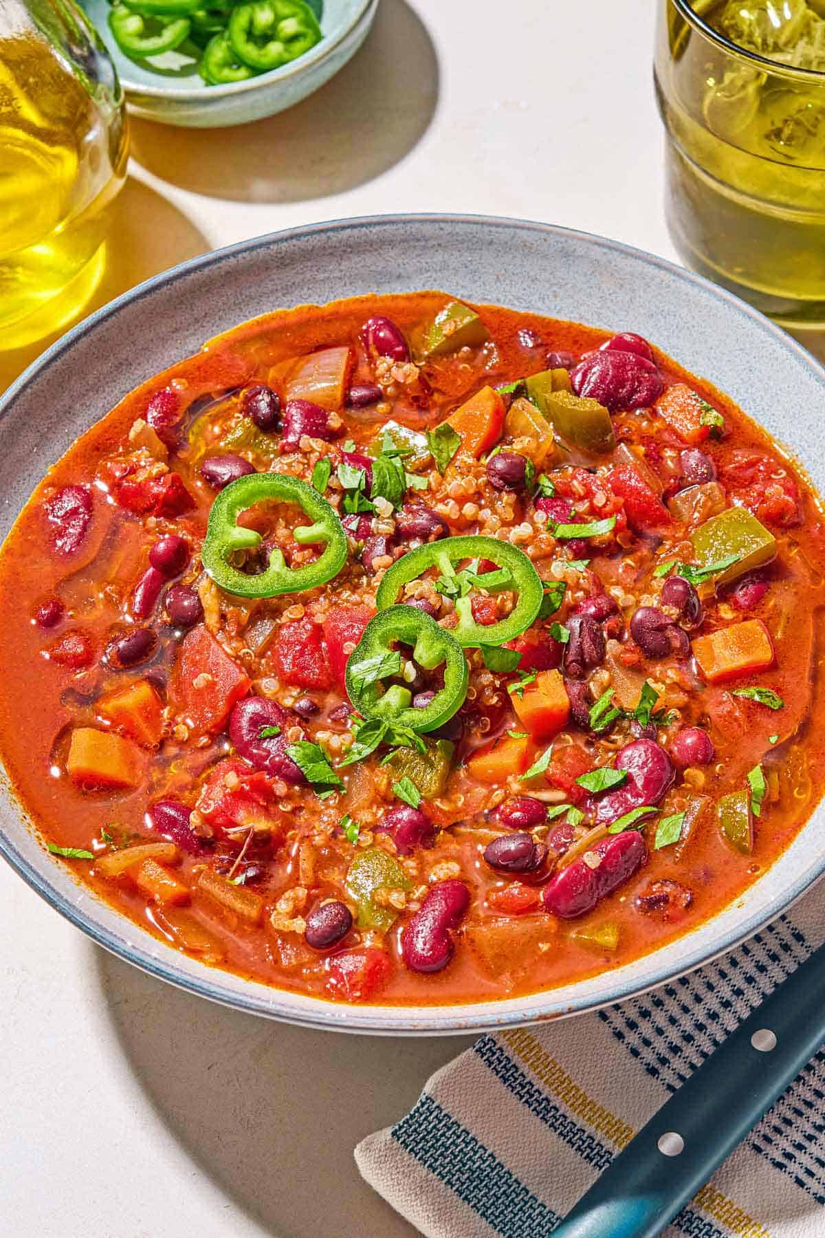 A bowl of vegan chili next to a cloth napkin with a spoon, a glass of water, a bowl of sliced jalapeños, and a bottle of olive oil.