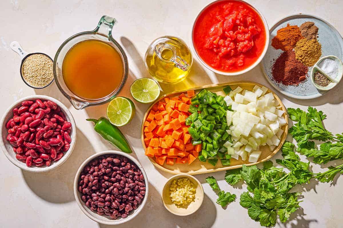 Ingredients for vegan chili including onions, green peppers, carrots, quinoa, olive oil, garlic, chopped tomatoes, broth, chili powder, paprika, cumin, allspice, salt, pepper, black beans, kidney beans, cilantro, parsley, lime and jalapeño.