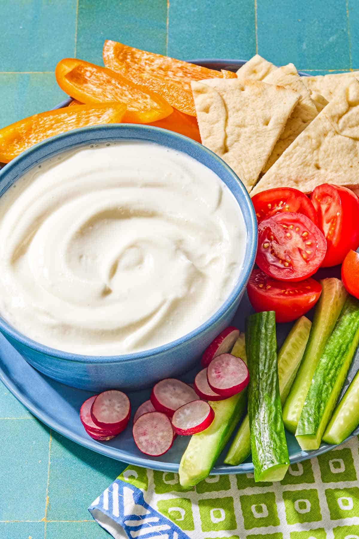 The toum recipe in a bowl on a platter with slices of orange bell pepper, tomato, radish, and cucumber along with pieces of pita bread. Next to this is a napkin.