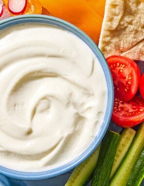The toum recipe in a bowl on a platter with slices of orange bell pepper, tomato, radish, and cucumber along with pieces of pita bread. Next to this is a napkin.