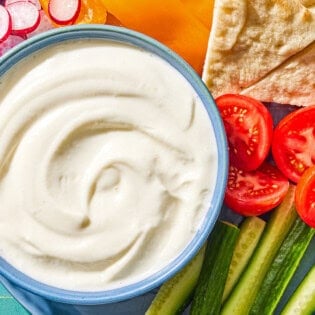 The toum recipe in a bowl on a platter with slices of orange bell pepper, tomato, radish, and cucumber along with pieces of pita bread. Next to this is a napkin.