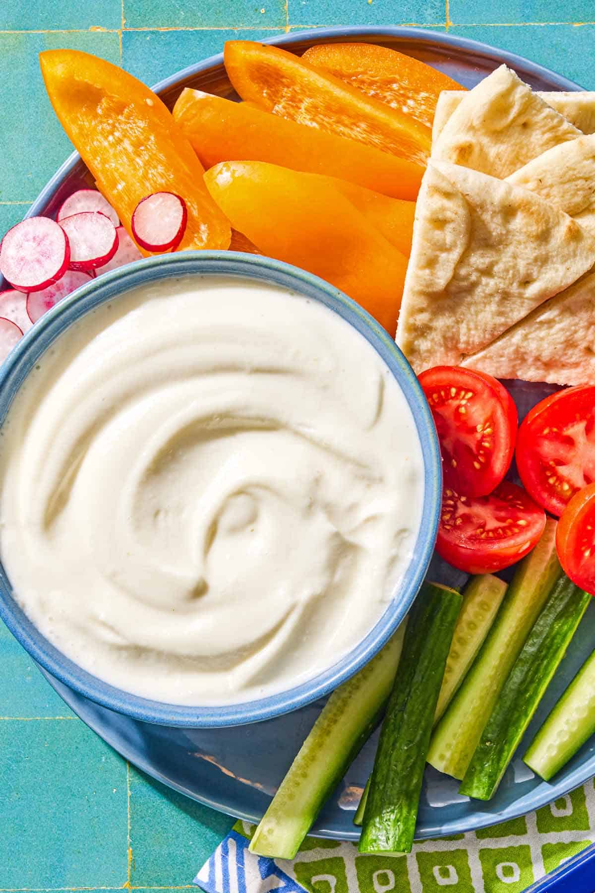 The toum recipe in a bowl on a platter with slices of orange bell pepper, tomato, radish, and cucumber along with pieces of pita bread. Next to this is a napkin.