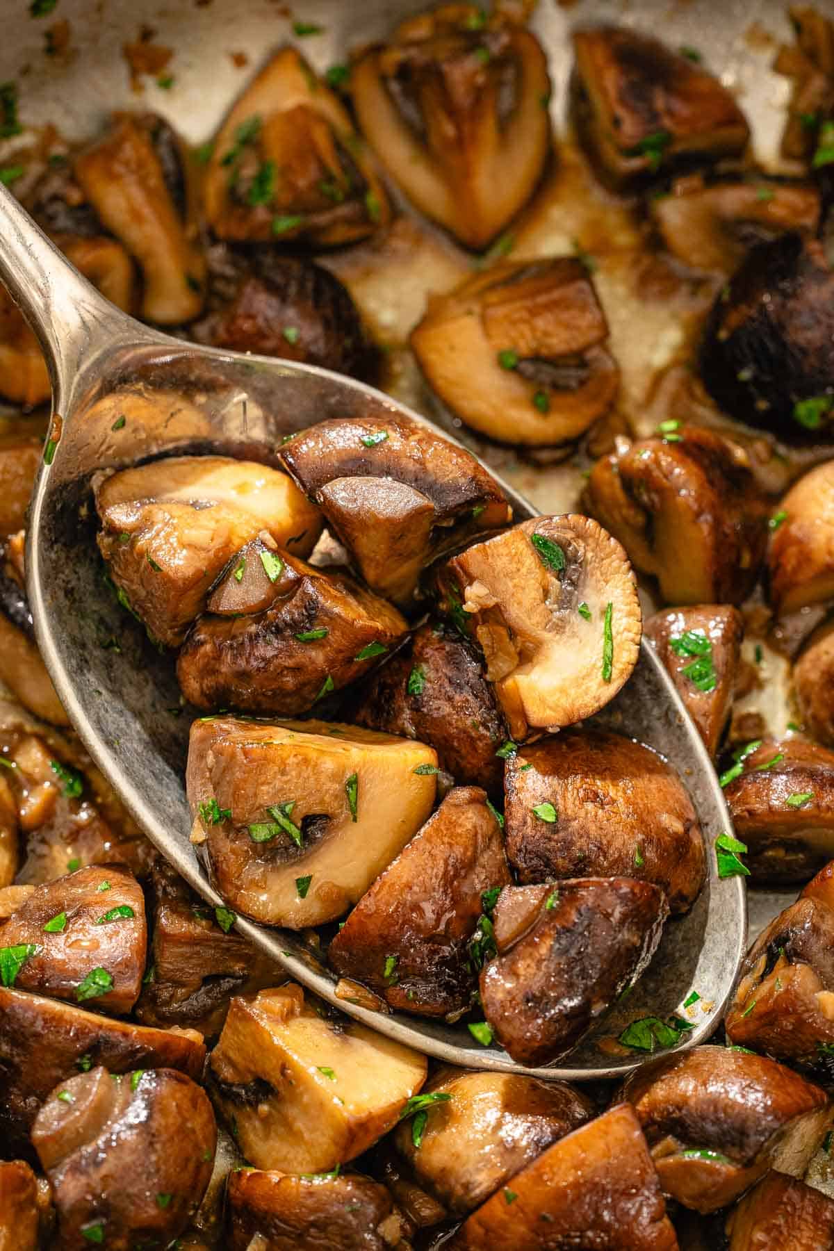 Spanish garlic mushrooms in a serving spoon being held over a bowl with the rest of the mushrooms.