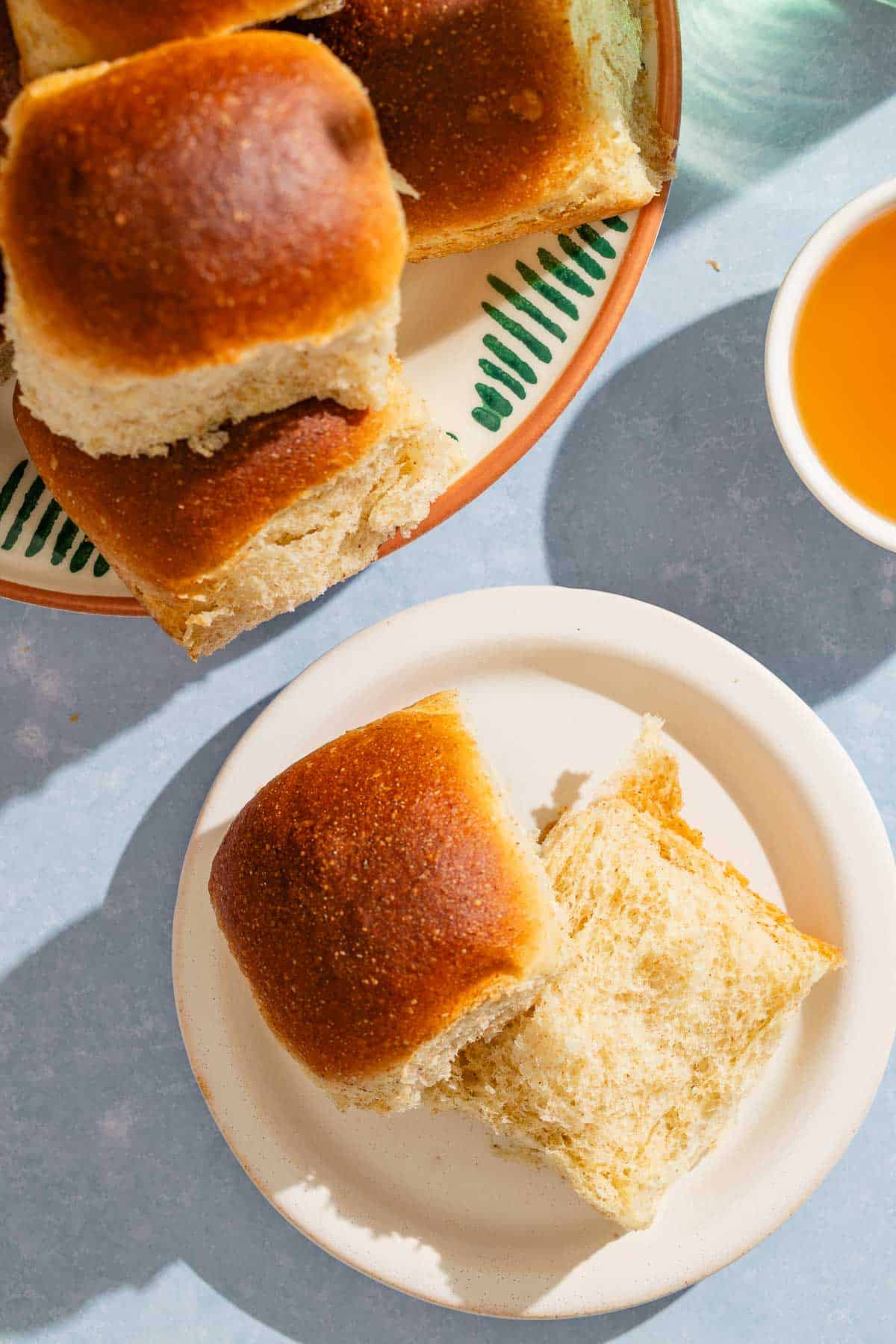 An overhead photo of a dinner roll on a plate split in half. Next to this is a stack of dinner rolls on a platter and a bowl of honey.