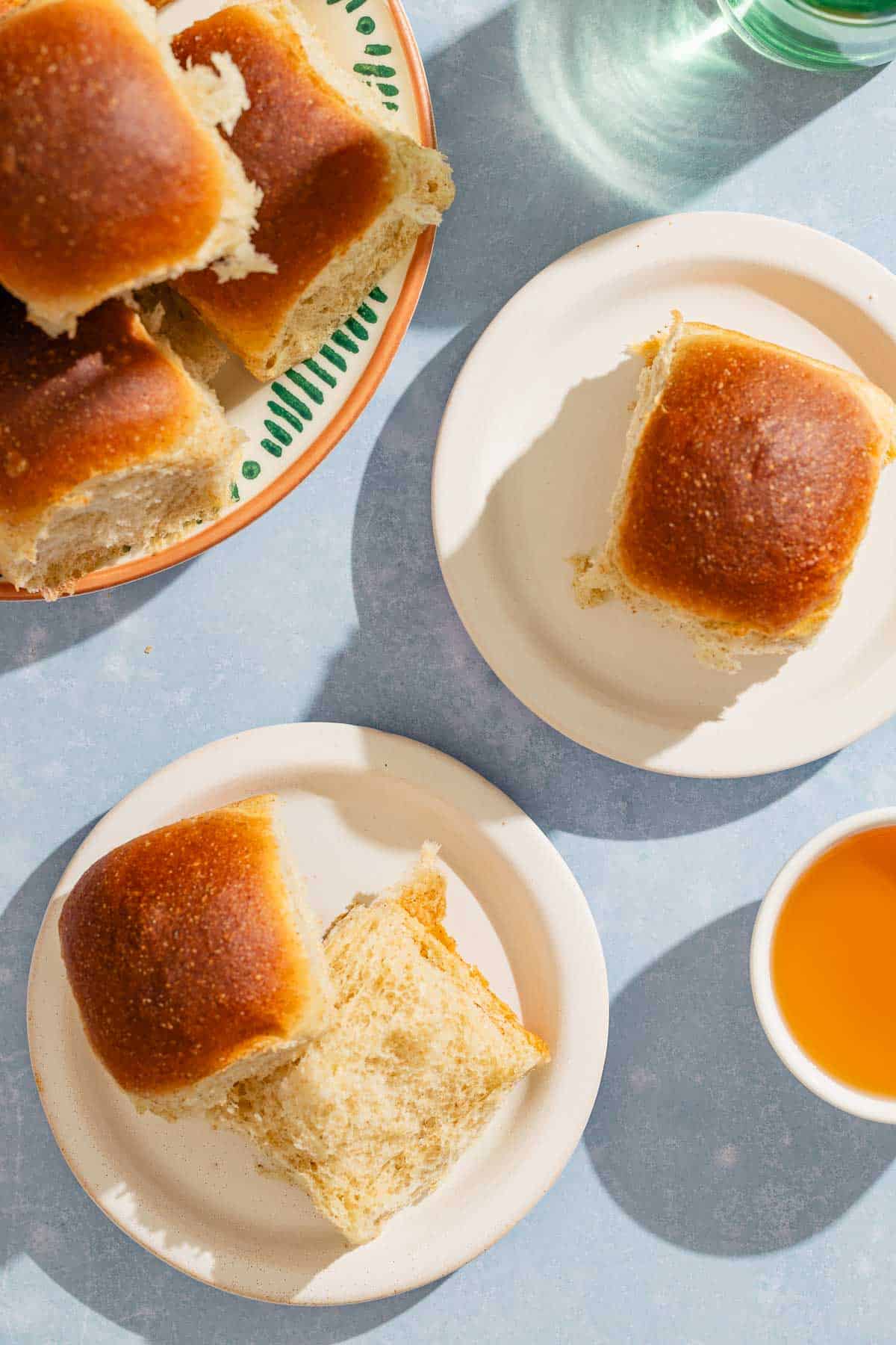 An overhead photo of two dinner rolls on two plates. Next to this is a stack of dinner rolls on a platter and a bowl of honey.