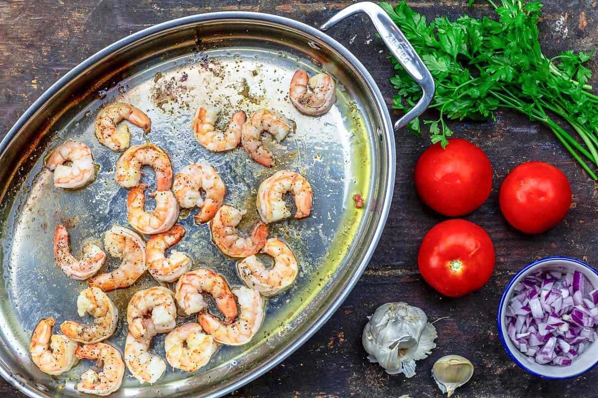 The shrimp for the shrimp pasta in a deep sided skillet after being sauteed. next to this is a bowl of chopped red onions, 3 tomatoes, parsley, and garlic.