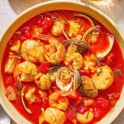 A serving of bouillabaisse in a bowl next to a spoon, a bowl of dill and a glass of white wine.