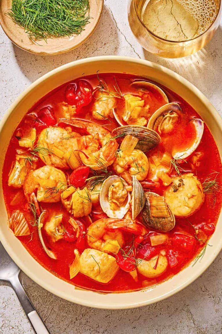 A serving of bouillabaisse in a bowl next to a spoon, a bowl of dill and a glass of white wine.