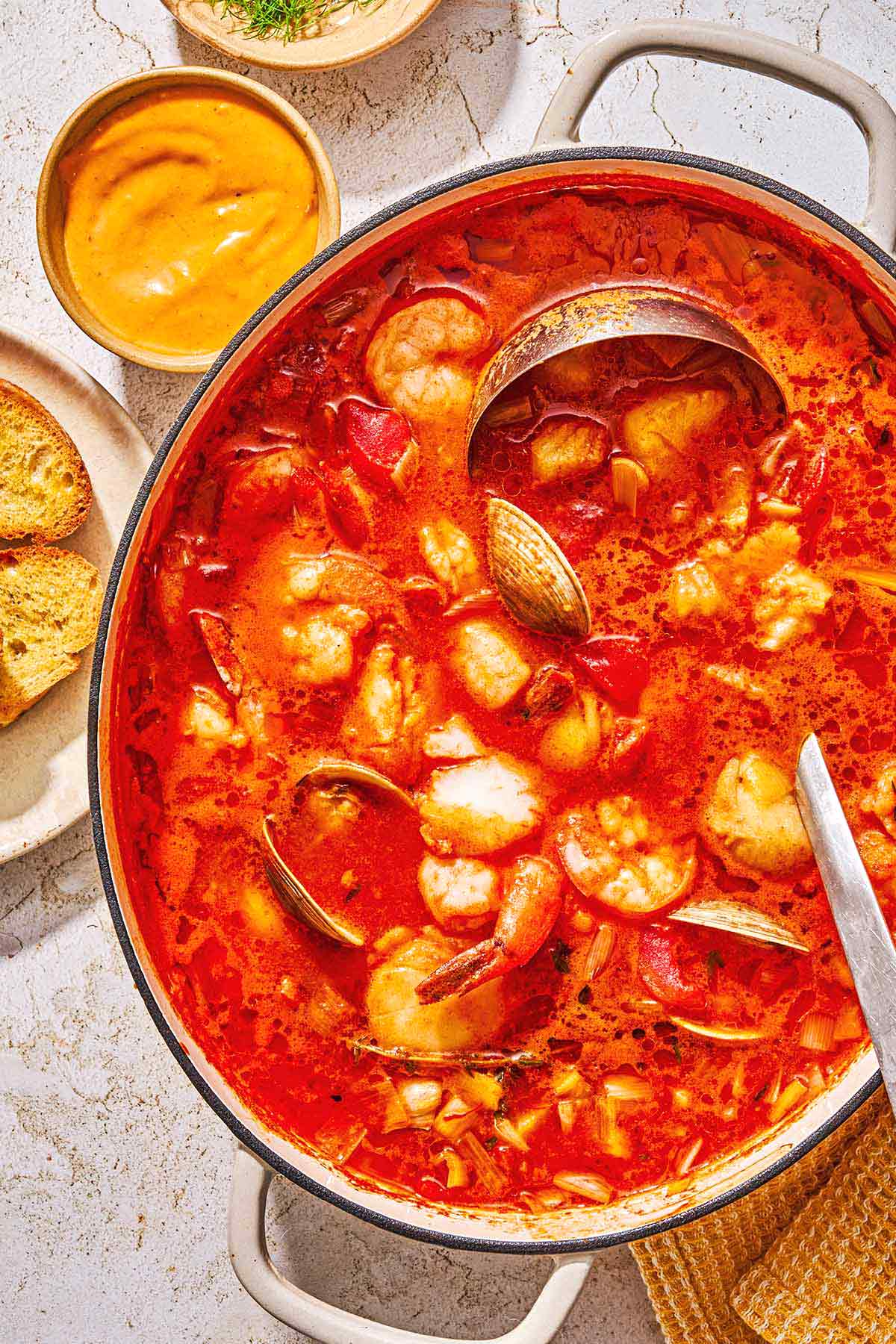 Bouillabaisse in a large pot with a ladle. Next to this is a cloth napkin, a bowl of the rouille, and slice of a French baguette.