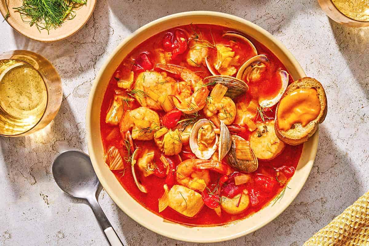 A serving of bouillabaisse in a bowl with a crostini. Next to this is a spoon, two glasses of white wine, and a bowl of dill.