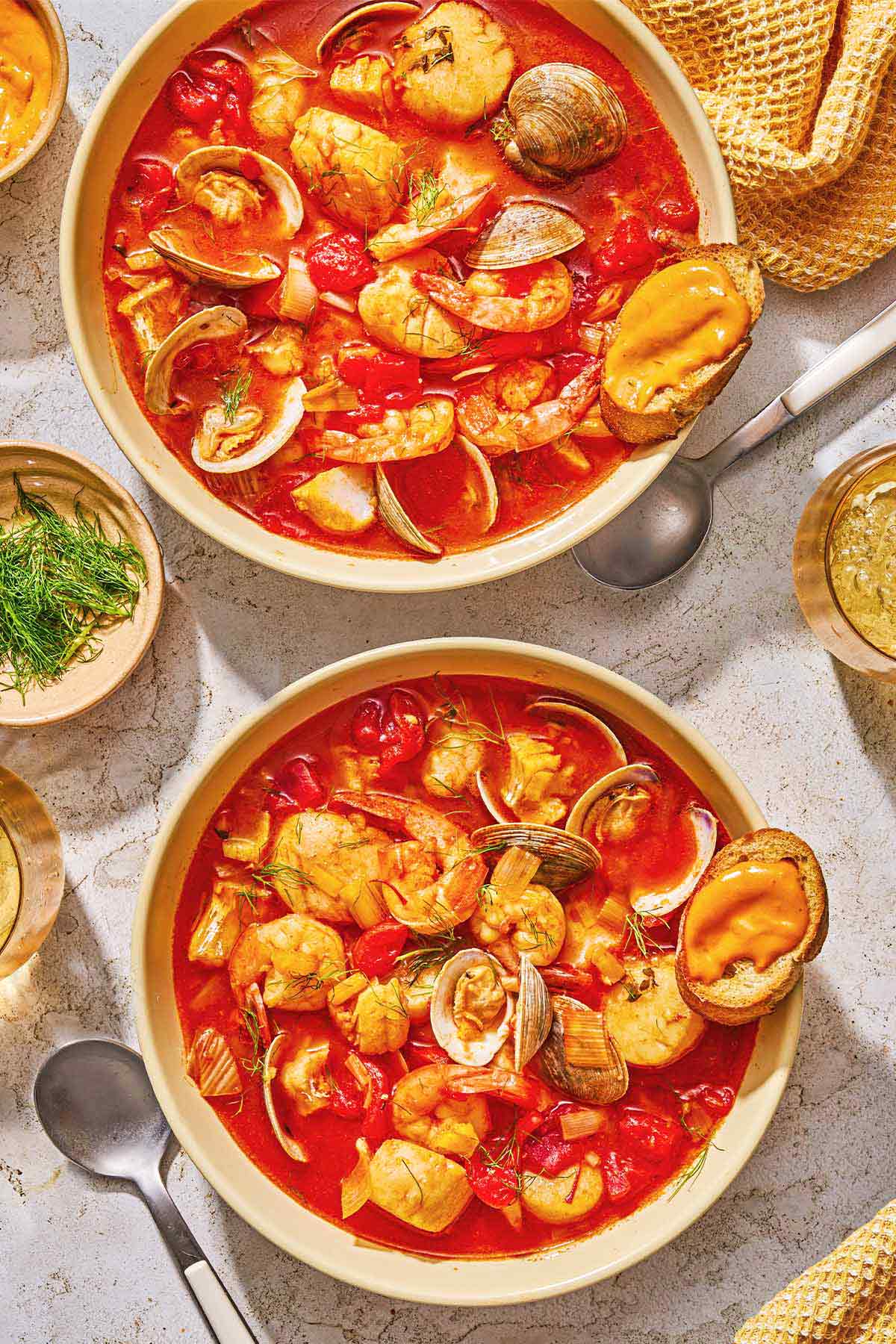 Two bowls of bouillabaisse each with a rouille topped crostini. Next to these are spoons, a cloth napkin, a glass of sparkling water and a bowl of dill.