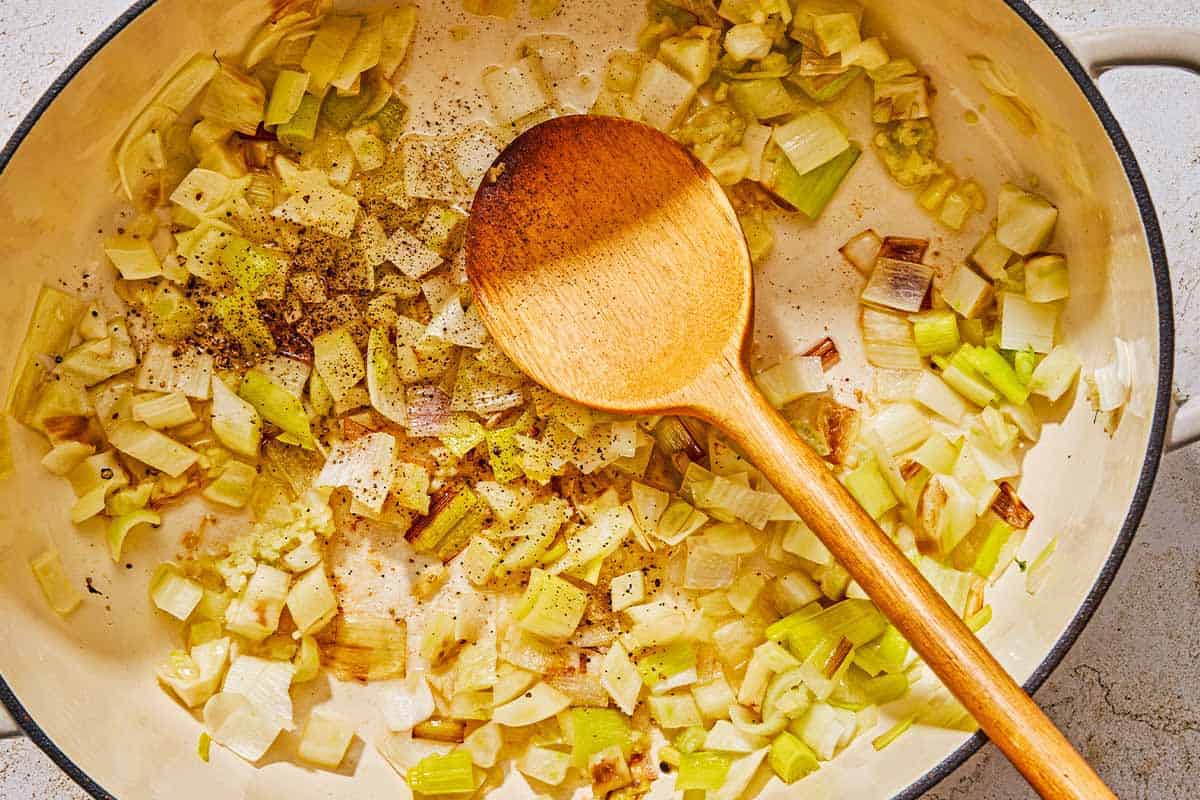 The leeks, fennel, garlic seasoned with salt and pepper being sautéed in a skillet with a wooden spoon.