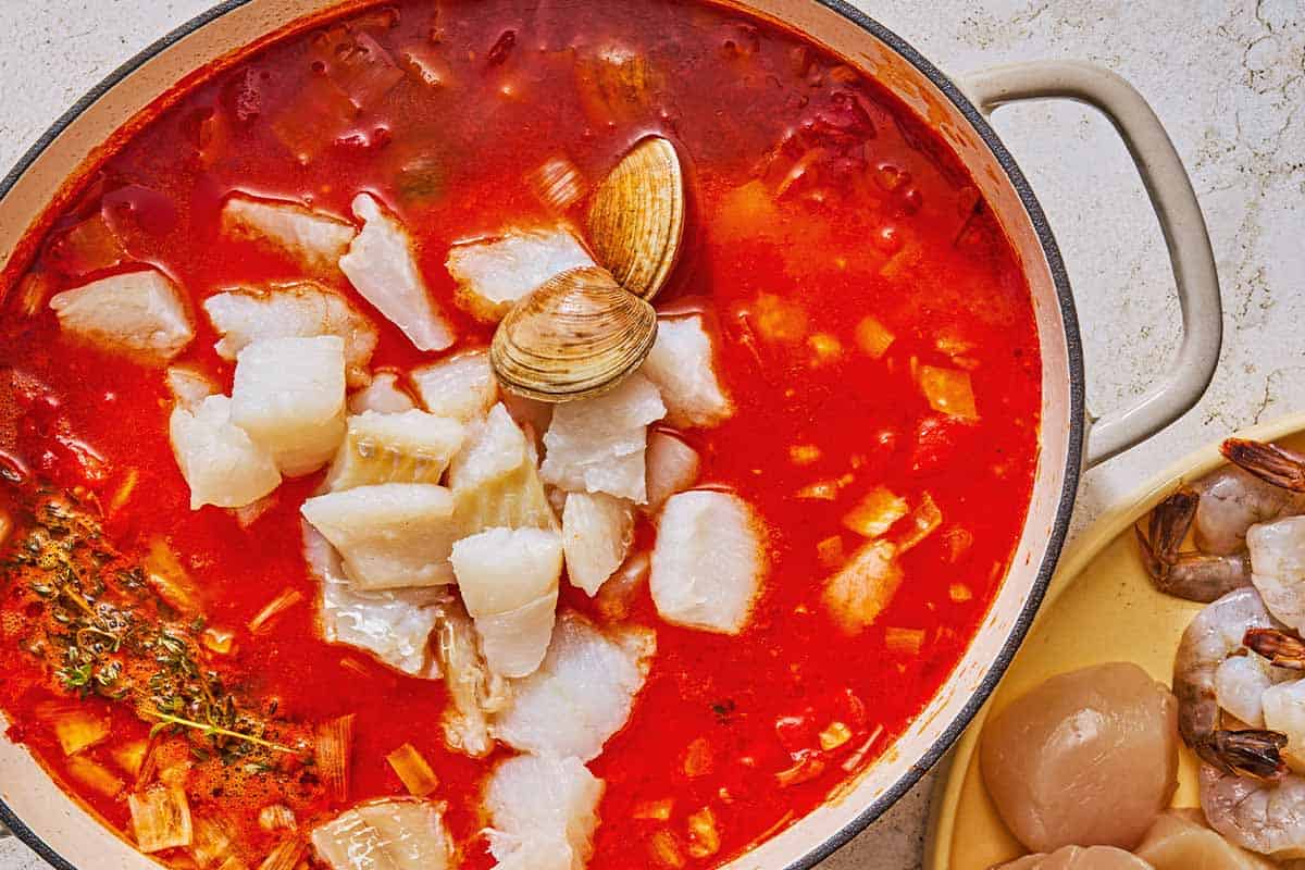 The tomato broth, vegetables, seasonings and herbs for the bouillabaisse in a pot along with the clams and white fish pieces. Next to this is a plate with uncooked shrimp and scallops.