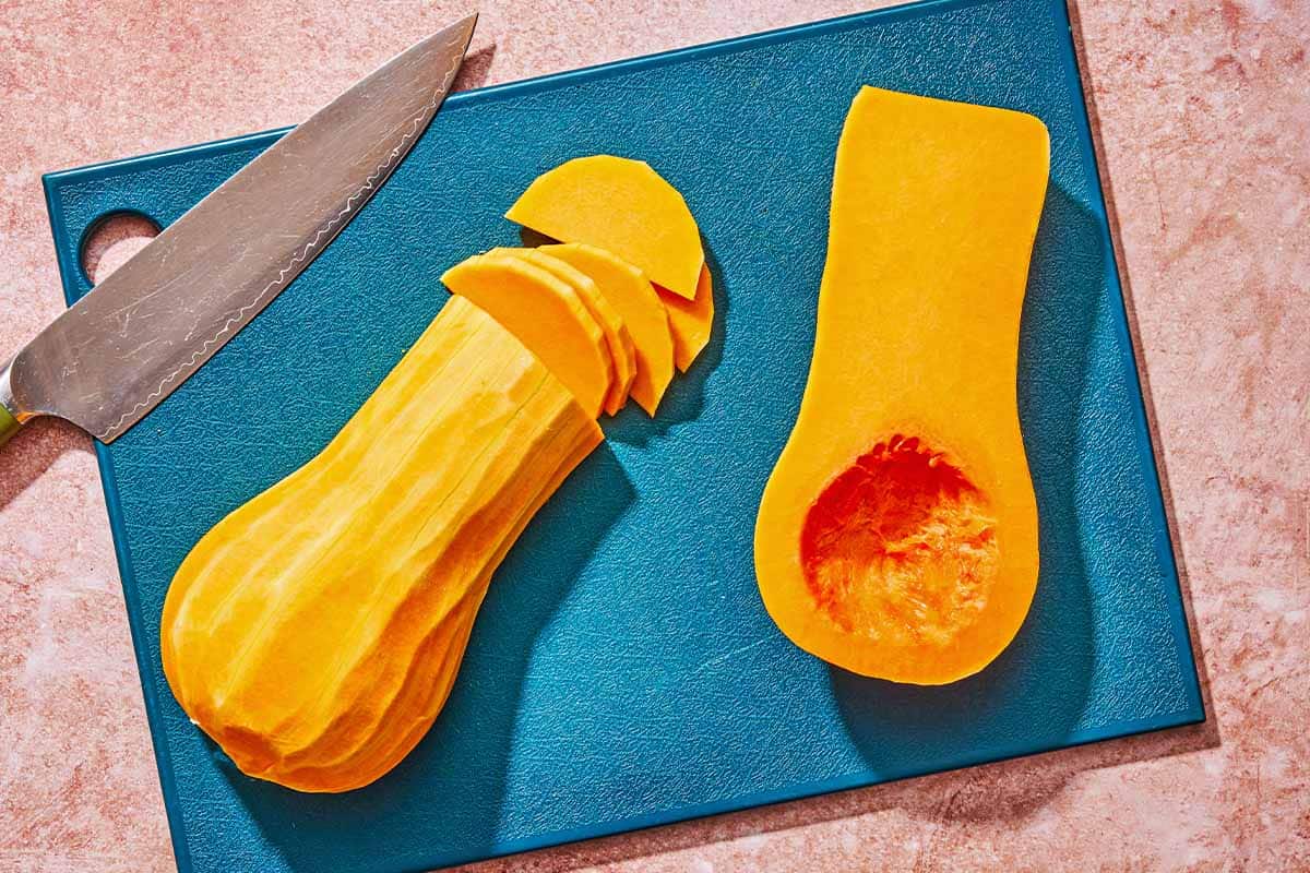 Two butternut squash halve on a cutting board with a knife. One of them is partially sliced.