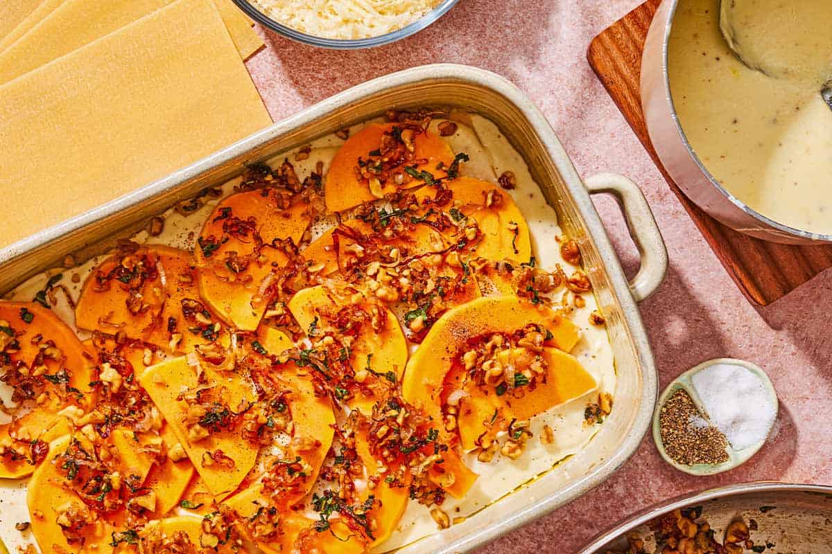 The partially assembled butternut squash lasagna in a baking dish surrounded by uncooked lasagna noodles, the cheese mixture in a bowl, the bechamel sauce in a saucepan and a bowl of salt and pepper.