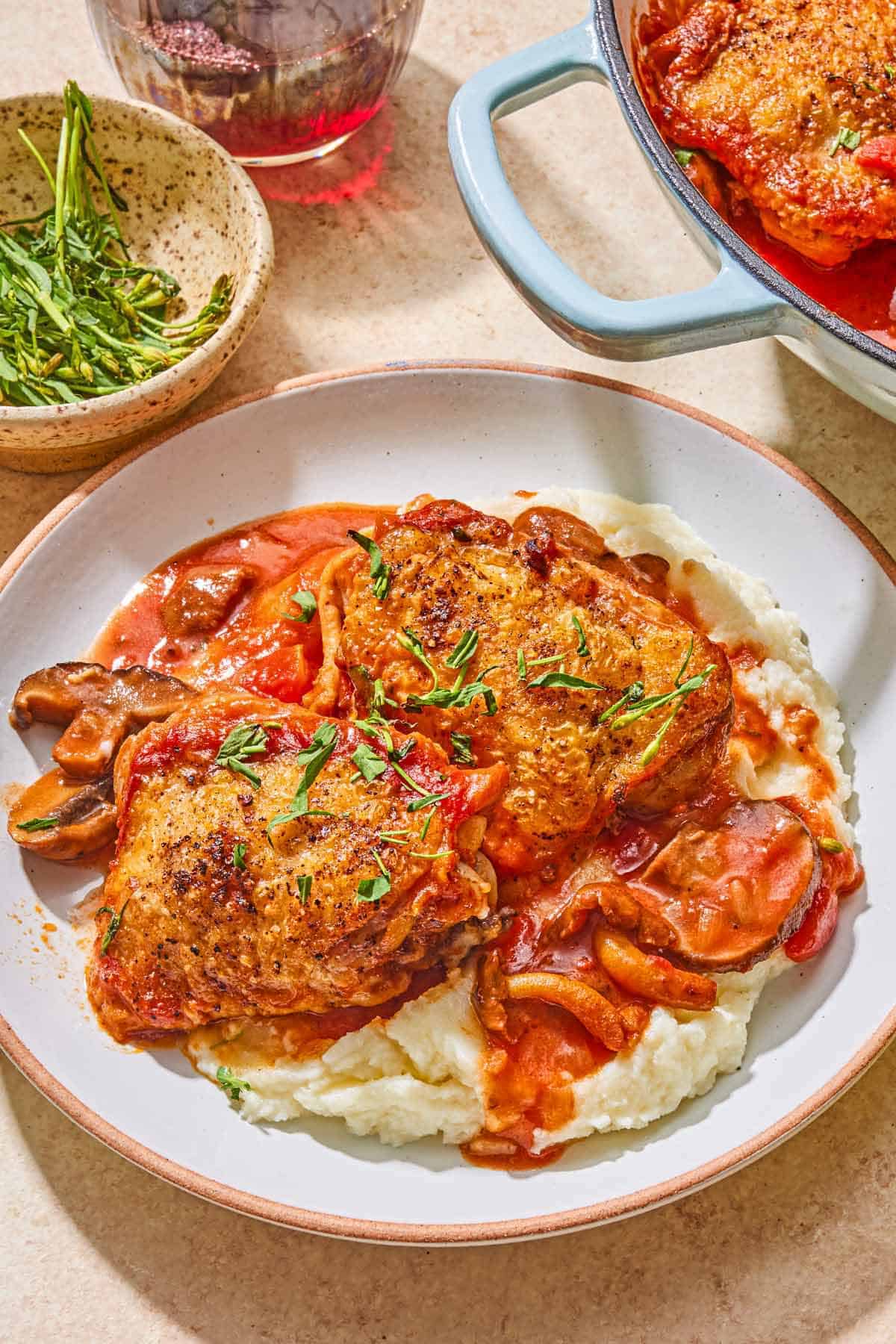 A serving of mashed potatoes topped with the chicken chasseur on a plate. Next to this is a bowl of herbs, a glass of white wine, and the rest of the chicken chasseur in a skillet.
