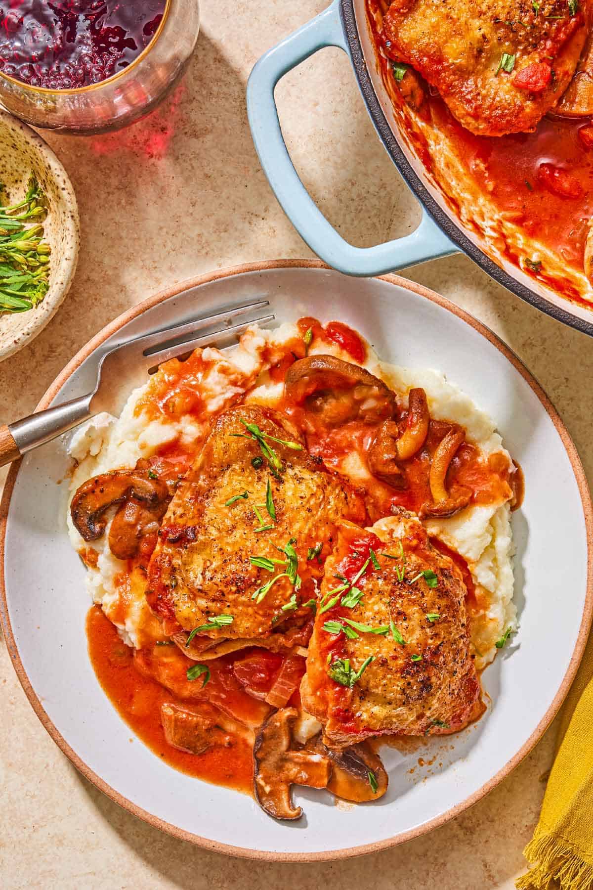 A serving of mashed potatoes topped with the chicken chasseur on a plate with a fork. Next to this is a bowl of herbs, a glass of white wine, and the rest of the chicken chasseur in a skillet.