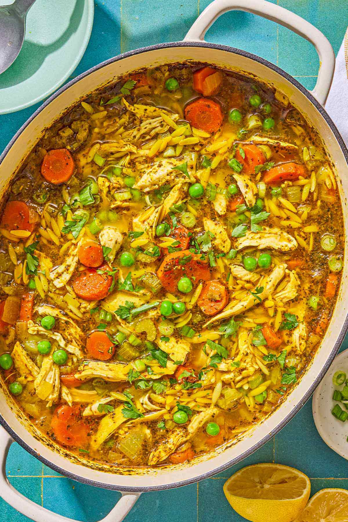 Chicken orzo soup in a pot. Next to this is a bowl of chopped green onions and 2 juiced lemon halves.