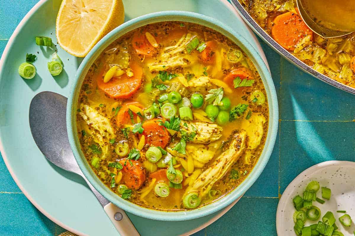 Chicken orzo soup in a bowl on a plate with a spoon and a juiced lemon half. Next to this is a bowl of chopped green onions and a pot with the rest of the soup and a ladle.