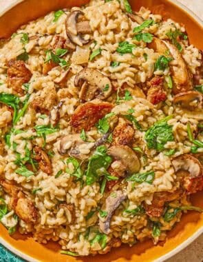 Chicken risotto in a serving bowl. Next to this is a a spoon, a cloth napkin and a bowls of grated Parmigiano Reggiano cheese, chopped parsley and pepper.