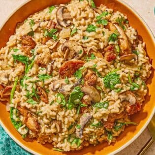 Chicken risotto in a serving bowl. Next to this is a a spoon, a cloth napkin and a bowls of grated Parmigiano Reggiano cheese, chopped parsley and pepper.