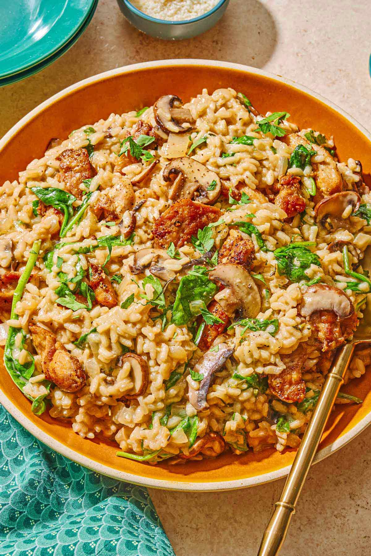 Chicken risotto in a serving bowl with a spoon. Next to this is a stack of 2 plates, a cloth napkin and a bowl of grated Parmigiano Reggiano cheese.
