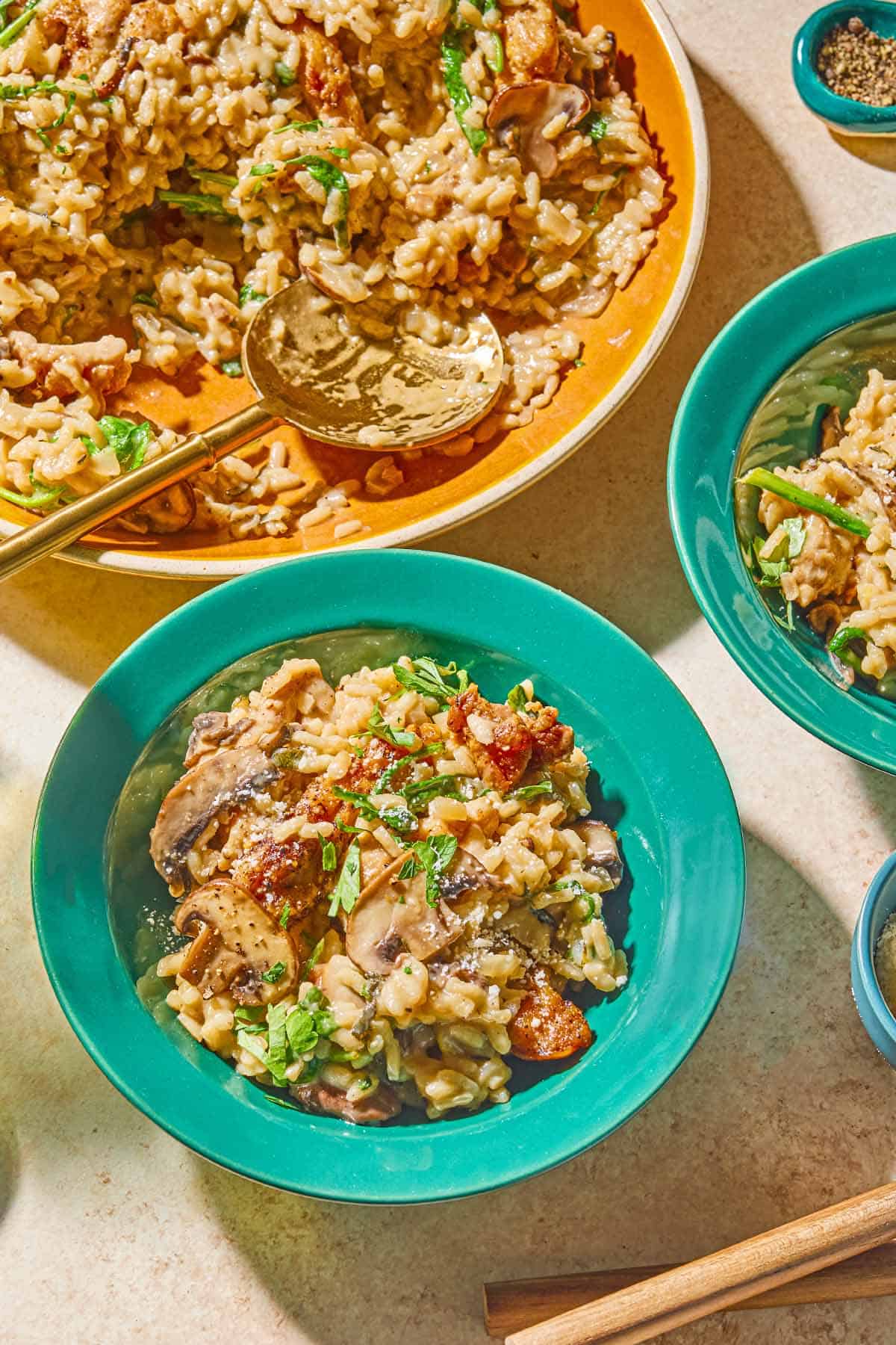 Two bowls of chicken risotto next to a serving bowl with the rest of the risotto and a serving spoon, and a bowl of pepper.