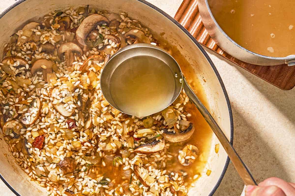 A ladle full of chicken stock being poured into the chicken risotto in a large pot. Next to this is a saucepan with the chicken stock on a trivet.