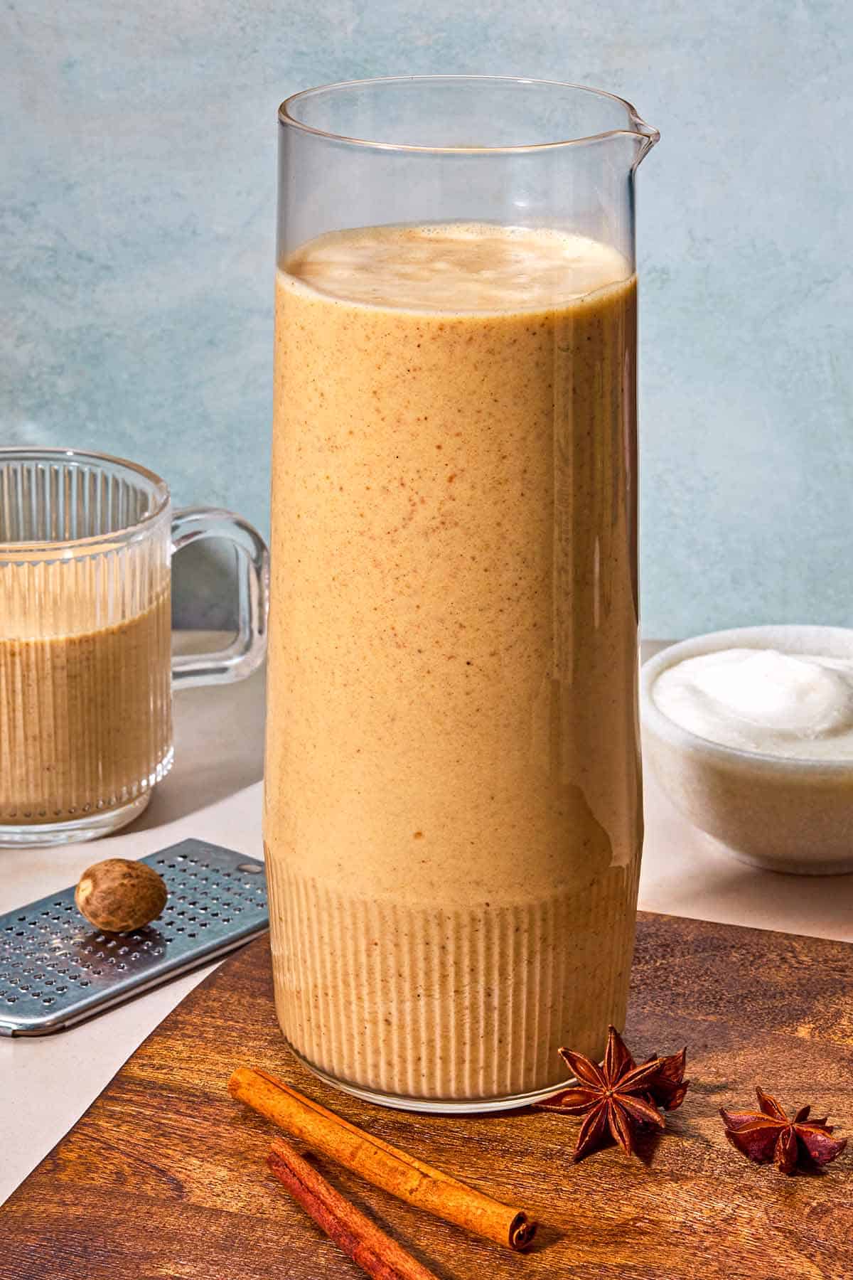 A pitcher of the vegan eggnog on a wooden tray with 2 cinnamon sticks and star anise. Next to this is a mug of the eggnog, a grater with a nutmeg and whipped coconut cream in a bowl.