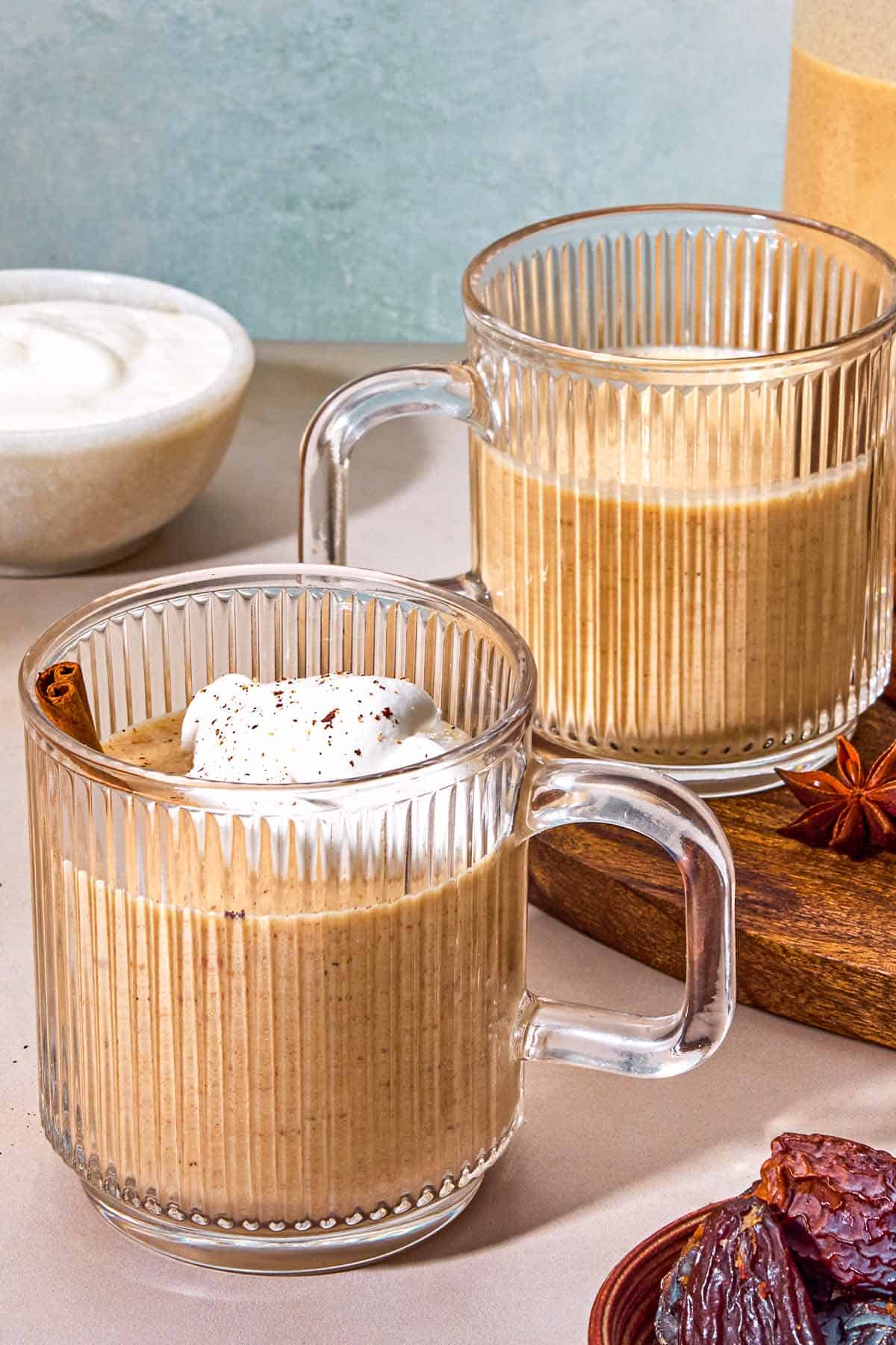 A mug of dairy-free eggnog garnished with a cinnamon stick and whipped coconut cream. Next to this is a bowl of dates, a bowl of whipped coconut cream, and a wooden tray with another mug of the eggnog and the rest of the eggnog in a pitcher.